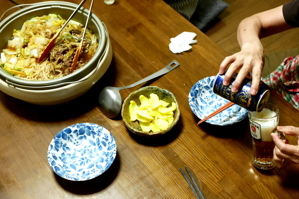 Sukiyaki pot on the table, an elder woman's hand is pouring a canned beer in a glass