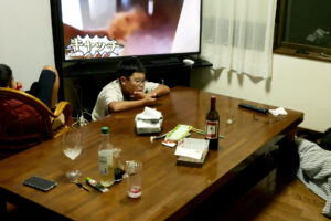 A child is using mobile phone at the table, another child is watching TV, there is a wooden table